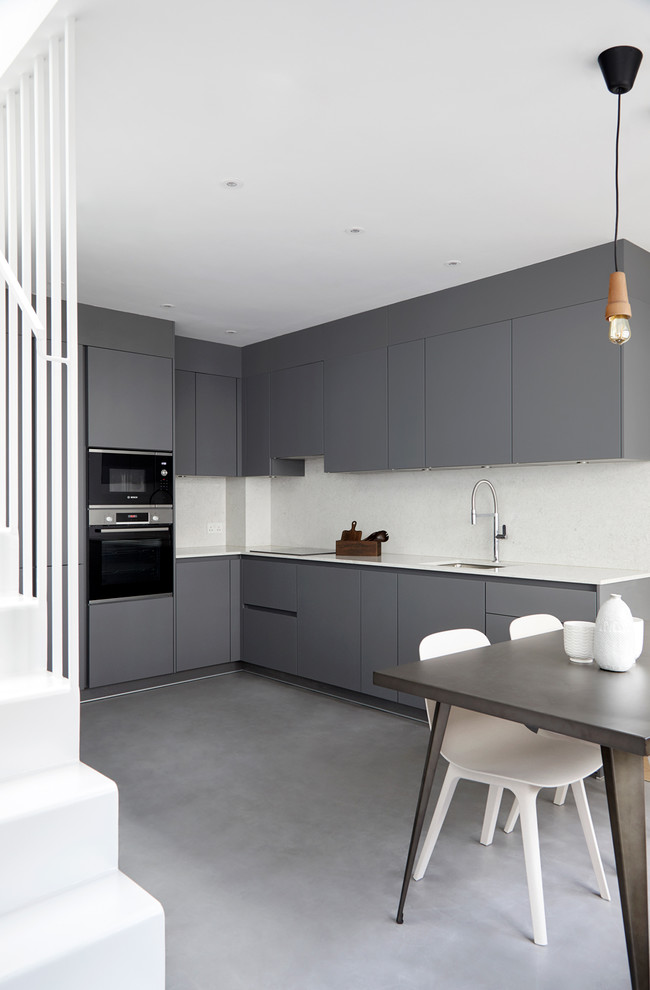 Moderne Wohnküche ohne Insel in L-Form mit Unterbauwaschbecken, flächenbündigen Schrankfronten, grauen Schränken, Küchenrückwand in Weiß, schwarzen Elektrogeräten, grauem Boden, weißer Arbeitsplatte und Betonboden in London