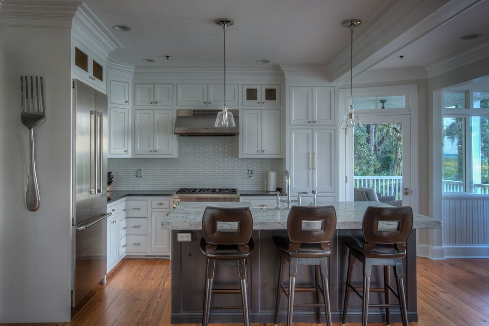 Mid-sized beach style l-shaped medium tone wood floor and brown floor eat-in kitchen photo in Atlanta with a farmhouse sink, recessed-panel cabinets, white cabinets, granite countertops, white backsplash, glass tile backsplash, stainless steel appliances, an island and black countertops
