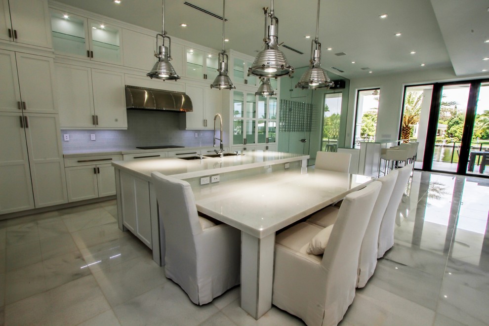 Large modern l-shaped kitchen in Miami with stainless steel appliances, grey splashback, glass tiled splashback, a submerged sink, shaker cabinets, white cabinets, engineered stone countertops, marble flooring, an island and white floors.