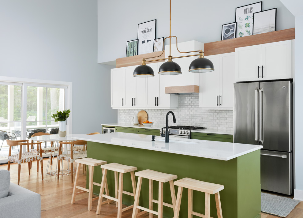 Coastal galley open plan kitchen in Chicago with a submerged sink, shaker cabinets, green cabinets, white splashback, mosaic tiled splashback, stainless steel appliances, medium hardwood flooring, an island, brown floors and white worktops.