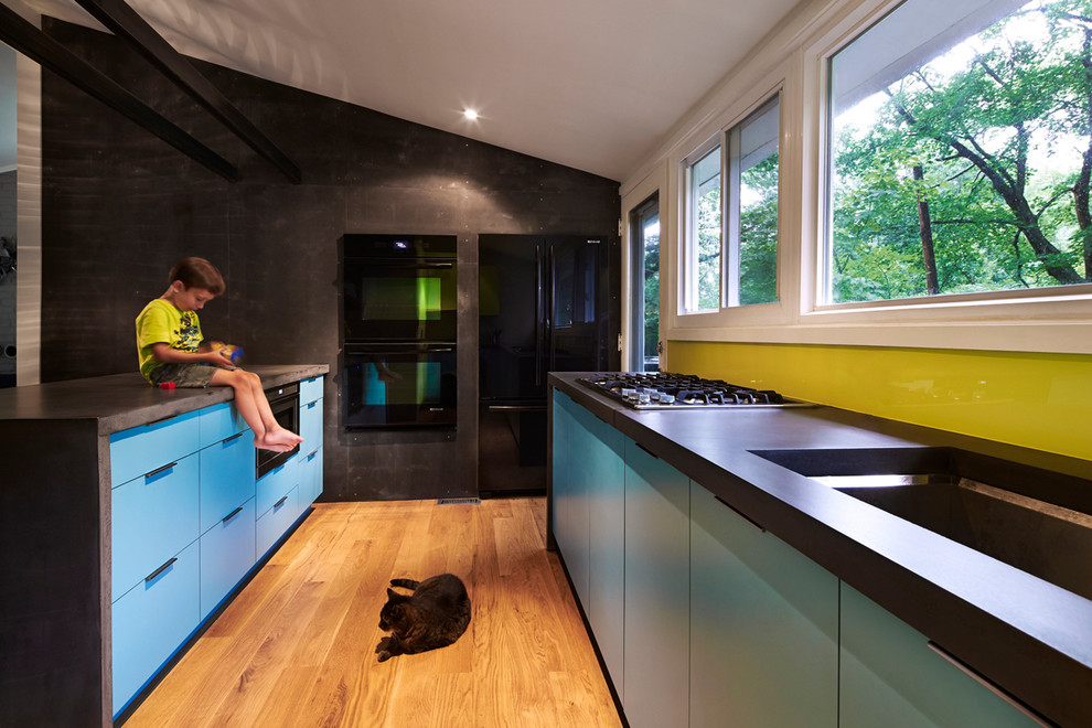 Foto på ett retro kök, med en undermonterad diskho, släta luckor, blå skåp, grönt stänkskydd, glaspanel som stänkskydd och svarta vitvaror