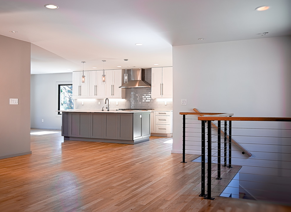Modern inredning av ett mellanstort vit vitt kök, med en undermonterad diskho, skåp i shakerstil, vita skåp, bänkskiva i kvartsit, grått stänkskydd, stänkskydd i tunnelbanekakel, rostfria vitvaror, ljust trägolv, en köksö och brunt golv