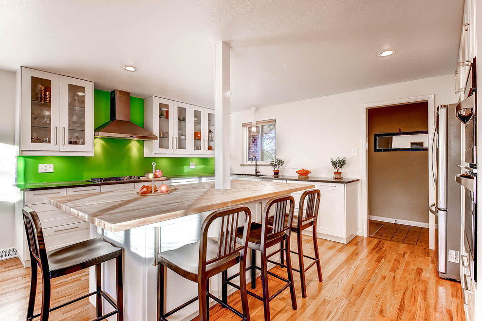 Foto på ett mellanstort 60 tals kök, med grönt stänkskydd, rostfria vitvaror, en enkel diskho, luckor med infälld panel, vita skåp, träbänkskiva, glaspanel som stänkskydd, mellanmörkt trägolv och en köksö