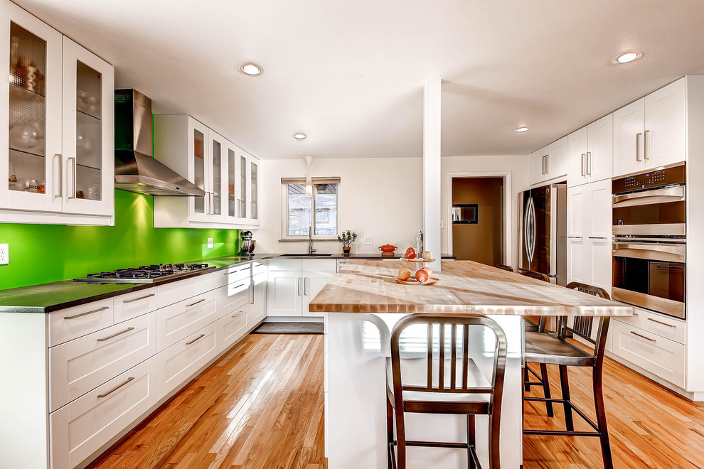 Mittelgroße Mid-Century Wohnküche in U-Form mit Küchenrückwand in Grün, Küchengeräten aus Edelstahl, Waschbecken, Schrankfronten mit vertiefter Füllung, weißen Schränken, Arbeitsplatte aus Holz, Glasrückwand, braunem Holzboden und Kücheninsel in Denver
