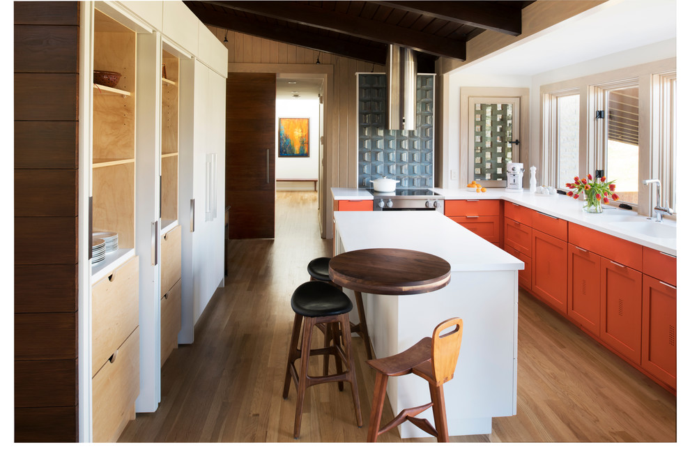 Mittelgroße Mid-Century Wohnküche in L-Form mit Doppelwaschbecken, Schrankfronten im Shaker-Stil, orangefarbenen Schränken, Mineralwerkstoff-Arbeitsplatte, Küchengeräten aus Edelstahl, hellem Holzboden und Kücheninsel in Atlanta