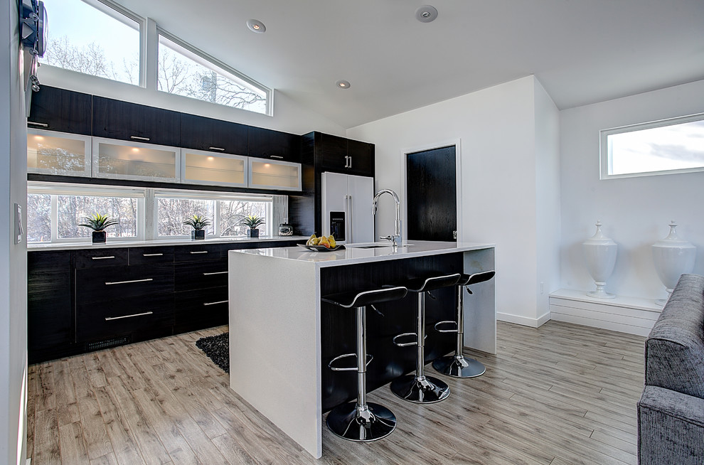 This is an example of a medium sized contemporary l-shaped open plan kitchen in Grand Rapids with a submerged sink, glass-front cabinets, engineered stone countertops, white splashback, glass tiled splashback, white appliances, light hardwood flooring, an island and black cabinets.