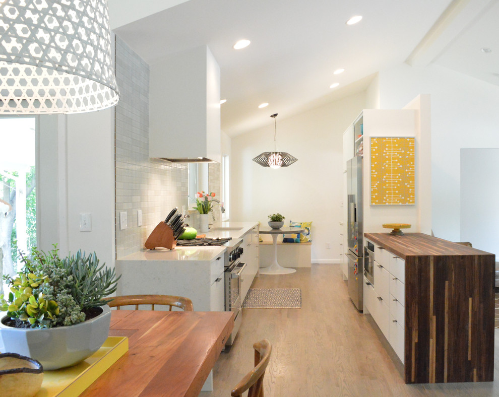 Example of a mid-sized mid-century modern galley dark wood floor and brown floor eat-in kitchen design in Denver with an undermount sink, flat-panel cabinets, white cabinets, solid surface countertops, gray backsplash, mosaic tile backsplash and stainless steel appliances