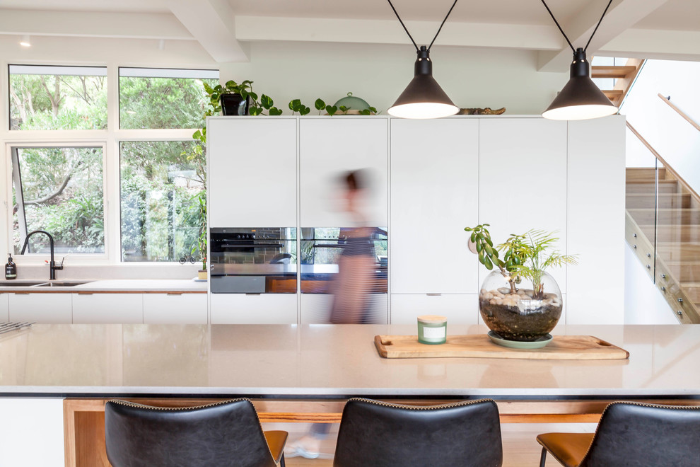 Inspiration for a large midcentury single-wall open plan kitchen in Melbourne with a submerged sink, white cabinets, engineered stone countertops, black appliances, medium hardwood flooring, an island, brown floors and white worktops.