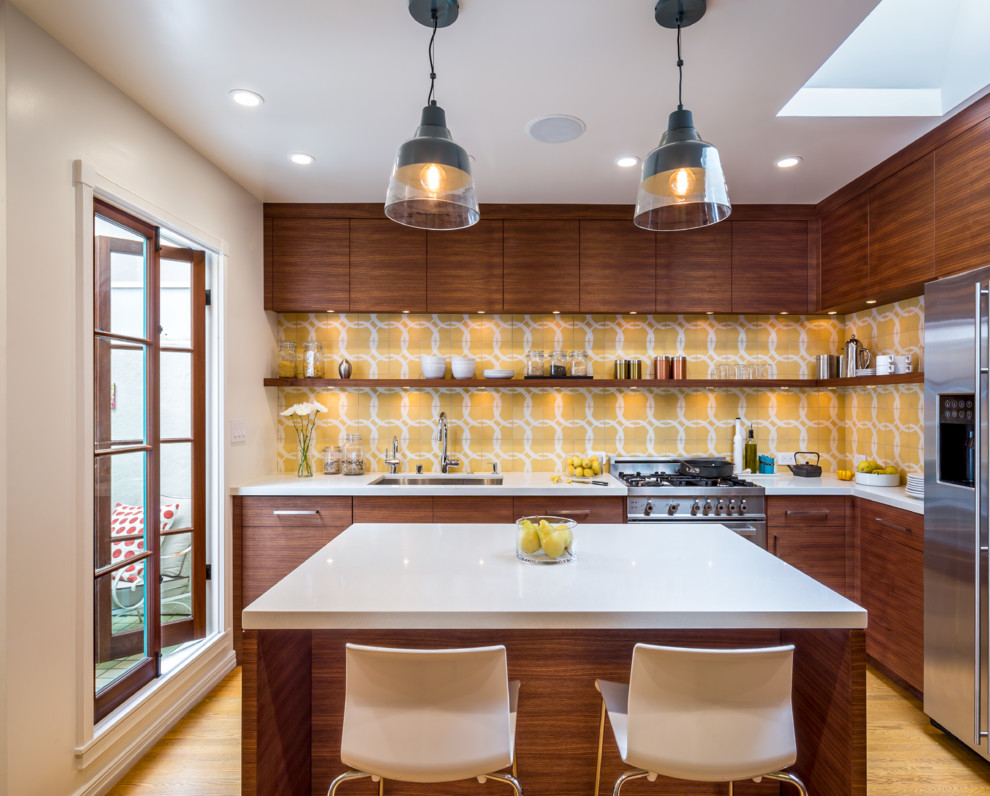 Foto på ett retro l-kök, med en undermonterad diskho, släta luckor, skåp i mellenmörkt trä, flerfärgad stänkskydd, rostfria vitvaror, ljust trägolv och en köksö