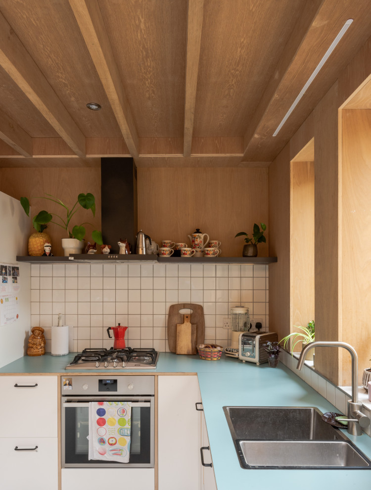 Photo of a retro l-shaped kitchen in London with wood worktops, blue worktops, a built-in sink, flat-panel cabinets, white cabinets, white splashback, stainless steel appliances and a wood ceiling.