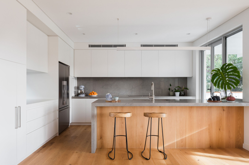 Photo of a large contemporary l-shaped open plan kitchen in Sydney with a submerged sink, white cabinets, engineered stone countertops, grey splashback, engineered quartz splashback, stainless steel appliances, medium hardwood flooring, an island, brown floors, grey worktops and flat-panel cabinets.