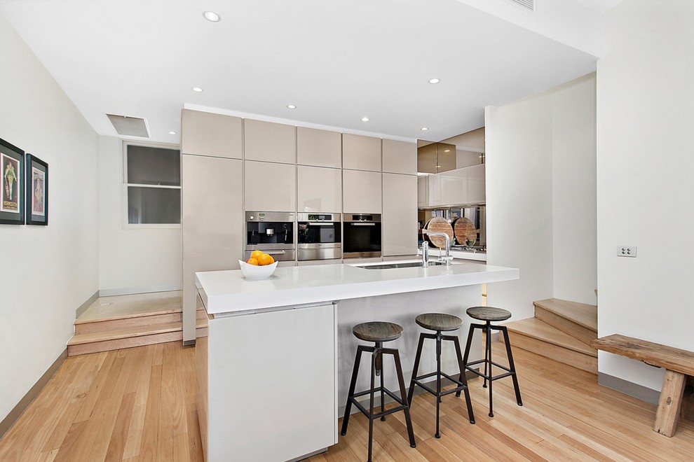 Contemporary galley open plan kitchen in Melbourne with a submerged sink, flat-panel cabinets, beige cabinets, stainless steel appliances, light hardwood flooring and an island.