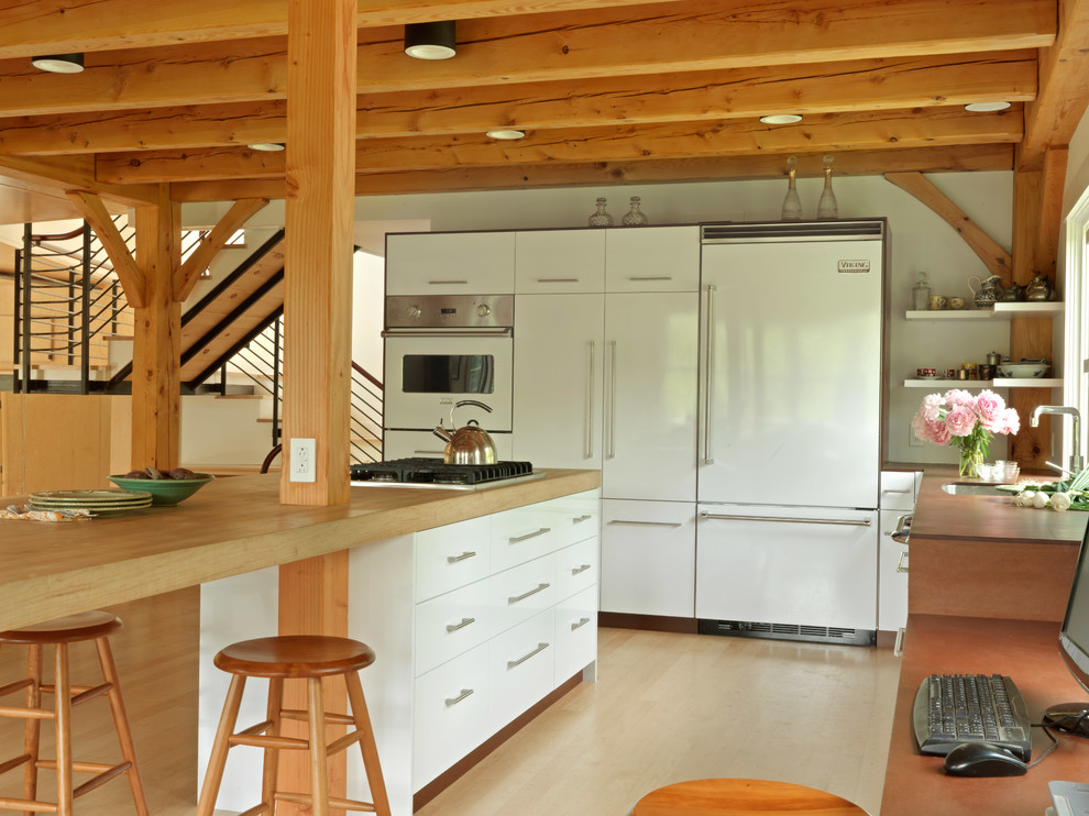 Rustik inredning av ett l-kök, med släta luckor, vita skåp, träbänkskiva, vita vitvaror, ljust trägolv och en köksö
