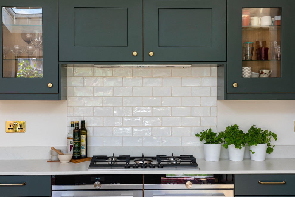 This is an example of a medium sized classic galley kitchen/diner in London with a built-in sink, shaker cabinets, green cabinets, granite worktops, white splashback, ceramic splashback, black appliances, light hardwood flooring, an island, brown floors and white worktops.