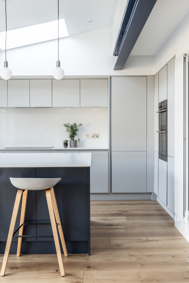 Mittelgroße Moderne Küche in L-Form mit flächenbündigen Schrankfronten, grauen Schränken, Küchenrückwand in Weiß, Elektrogeräten mit Frontblende, Kücheninsel, beigem Boden und weißer Arbeitsplatte in West Midlands