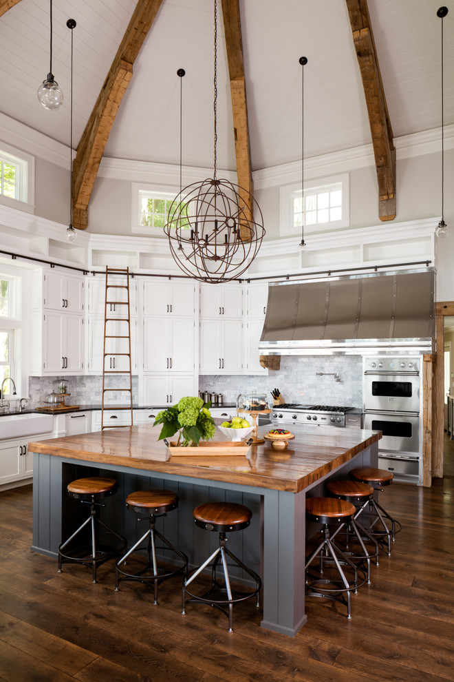 Bild på ett mycket stort lantligt l-kök, med en rustik diskho, skåp i shakerstil, vita skåp, träbänkskiva, grått stänkskydd, rostfria vitvaror, mörkt trägolv och en köksö