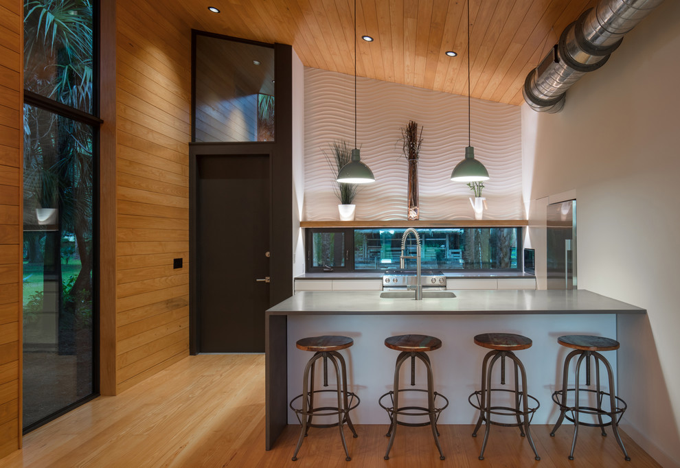 Inredning av ett modernt litet grå linjärt grått kök med öppen planlösning, med en integrerad diskho, släta luckor, vita skåp, bänkskiva i betong, fönster som stänkskydd, rostfria vitvaror, ljust trägolv, en halv köksö och flerfärgat golv