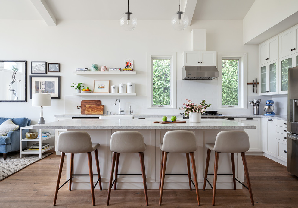 Réalisation d'une cuisine ouverte tradition en L avec un placard à porte shaker, des portes de placard blanches, plan de travail en marbre, un électroménager en acier inoxydable, un sol en bois brun, îlot, un plan de travail blanc, un évier de ferme, une crédence blanche et un sol marron.