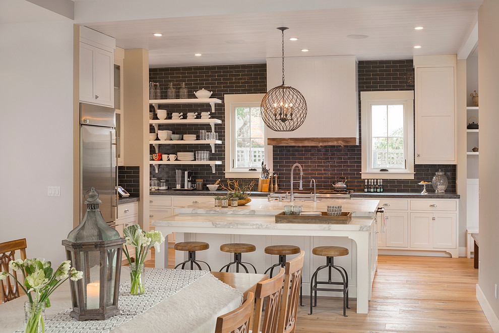 Foto de cocina de estilo de casa de campo con armarios estilo shaker, puertas de armario blancas, salpicadero negro, salpicadero de azulejos tipo metro, electrodomésticos de acero inoxidable, suelo de madera en tonos medios y una isla