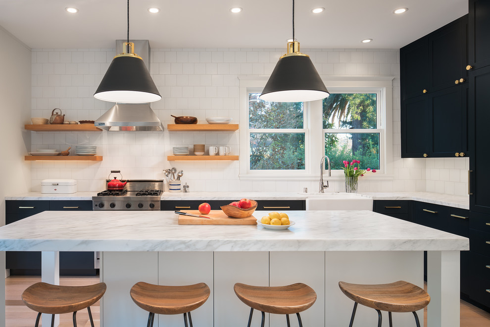 Traditional l-shaped kitchen in San Francisco with a belfast sink, shaker cabinets, black cabinets, white splashback, stainless steel appliances, light hardwood flooring, an island and beige floors.