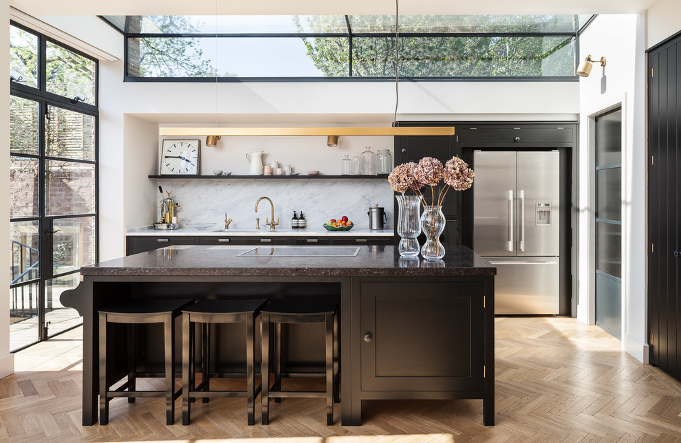 Idée de décoration pour une cuisine parallèle tradition de taille moyenne avec un placard à porte shaker, des portes de placard noires, une crédence grise, un électroménager en acier inoxydable, parquet clair, îlot et un sol beige.