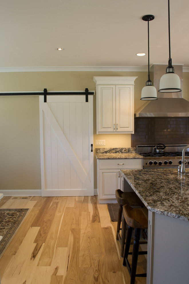 Klassisk inredning av ett stort kök, med en undermonterad diskho, luckor med upphöjd panel, vita skåp, granitbänkskiva, svart stänkskydd, stänkskydd i tunnelbanekakel, rostfria vitvaror, ljust trägolv och en köksö
