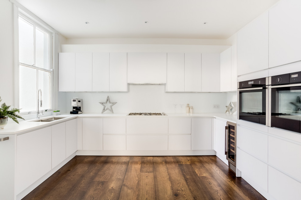 Modern inredning av ett vit vitt kök, med vita skåp, vitt stänkskydd, glaspanel som stänkskydd, mörkt trägolv och brunt golv