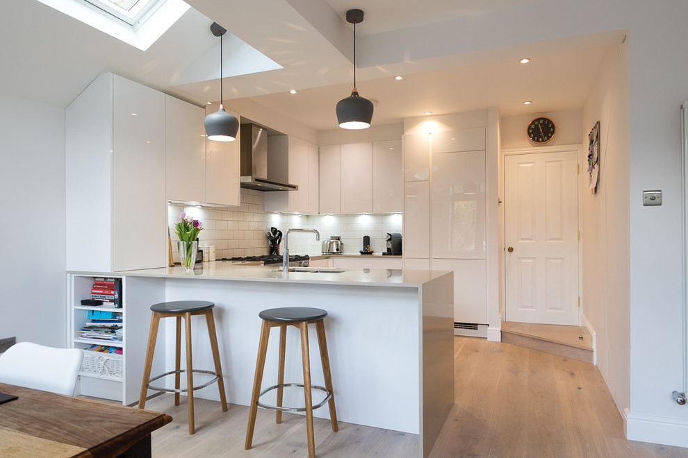 Photo of a small contemporary u-shaped kitchen in Surrey with flat-panel cabinets, white cabinets, quartz worktops, white splashback, ceramic splashback, stainless steel appliances, light hardwood flooring, a breakfast bar, brown floors, a submerged sink and white worktops.