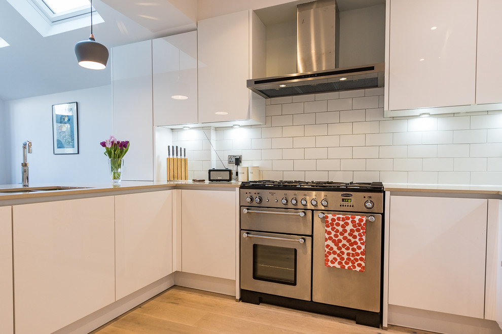 Photo of a small contemporary u-shaped open plan kitchen in Surrey with a built-in sink, flat-panel cabinets, white cabinets, quartz worktops, white splashback, ceramic splashback, stainless steel appliances, light hardwood flooring, a breakfast bar and brown floors.