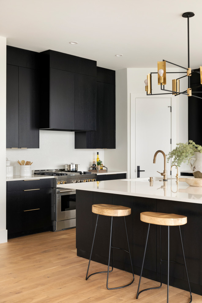 Inspiration for a large contemporary l-shaped kitchen/diner in Minneapolis with a belfast sink, flat-panel cabinets, black cabinets, granite worktops, white splashback, ceramic splashback, stainless steel appliances, light hardwood flooring, an island, brown floors and white worktops.