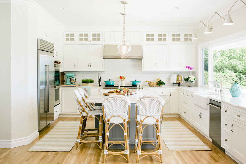 This is an example of a coastal l-shaped enclosed kitchen in Los Angeles with a belfast sink, shaker cabinets, white cabinets, white splashback, metro tiled splashback, stainless steel appliances, light hardwood flooring, an island and brown floors.