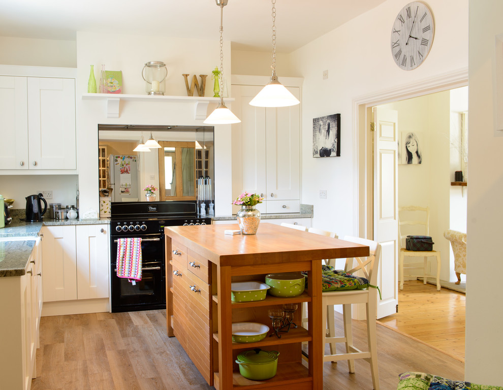 This is an example of a farmhouse l-shaped kitchen/diner in Kent with a belfast sink, shaker cabinets, white cabinets, granite worktops, mirror splashback, an island and black appliances.