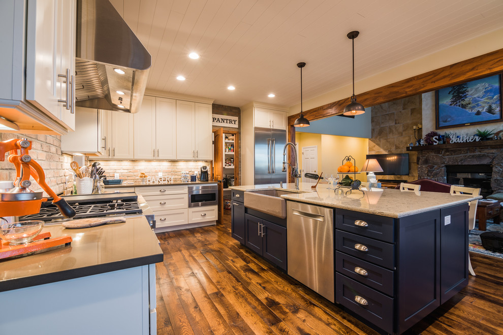 This is an example of a medium sized country galley kitchen/diner in Denver with a belfast sink, shaker cabinets, white cabinets, engineered stone countertops, grey splashback, brick splashback, stainless steel appliances, medium hardwood flooring, an island, brown floors and grey worktops.