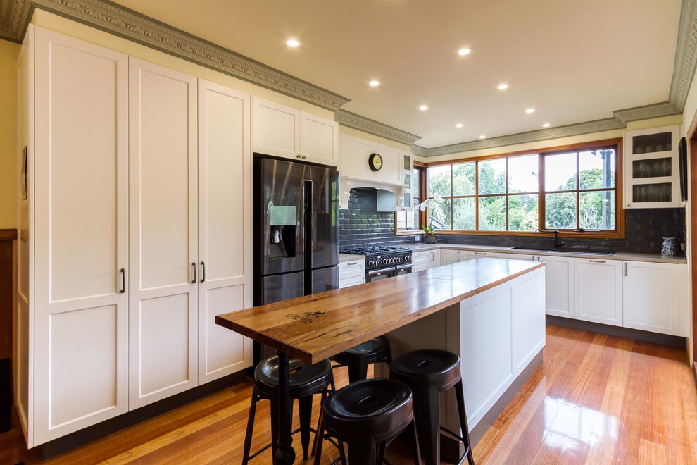 Example of a large transitional l-shaped medium tone wood floor eat-in kitchen design in Melbourne with shaker cabinets, gray backsplash, subway tile backsplash, a drop-in sink, white cabinets, black appliances and an island