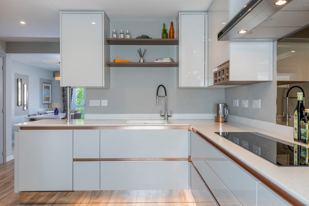 This is an example of a medium sized contemporary l-shaped open plan kitchen in Buckinghamshire with a single-bowl sink, flat-panel cabinets, grey cabinets, composite countertops, metallic splashback, glass sheet splashback, stainless steel appliances, vinyl flooring, no island, brown floors and white worktops.
