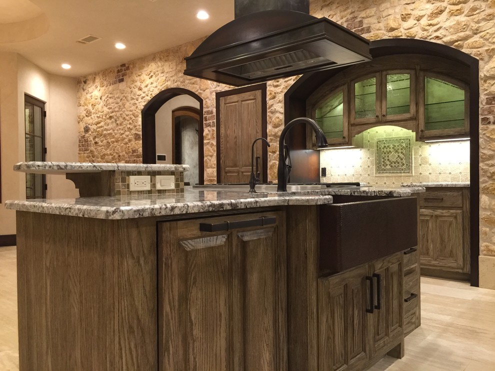 Mid-sized tuscan travertine floor eat-in kitchen photo in Austin with a farmhouse sink, raised-panel cabinets, distressed cabinets, granite countertops, beige backsplash, stone tile backsplash, stainless steel appliances and an island