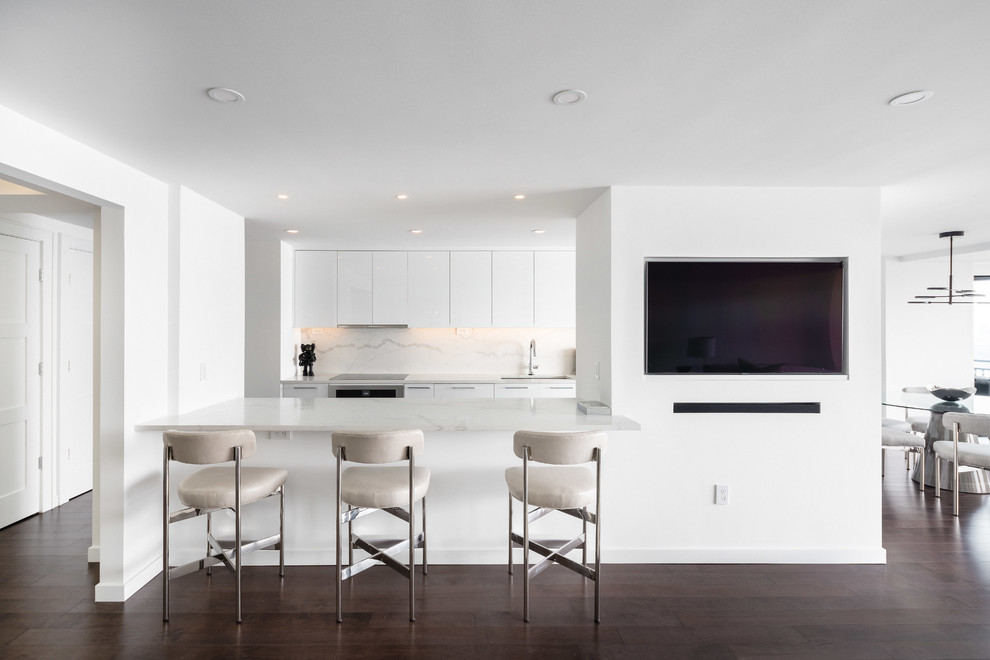Inredning av ett modernt vit vitt kök med öppen planlösning, med en undermonterad diskho, släta luckor, vita skåp, vitt stänkskydd, stänkskydd i sten, mörkt trägolv, brunt golv, bänkskiva i kvarts och rostfria vitvaror