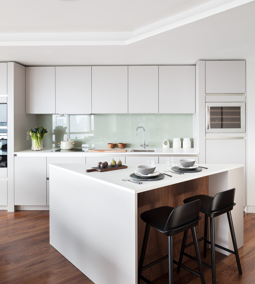 Inspiration for a medium sized modern single-wall open plan kitchen in London with a built-in sink, flat-panel cabinets, white cabinets, composite countertops, white splashback, glass sheet splashback, integrated appliances, dark hardwood flooring and an island.