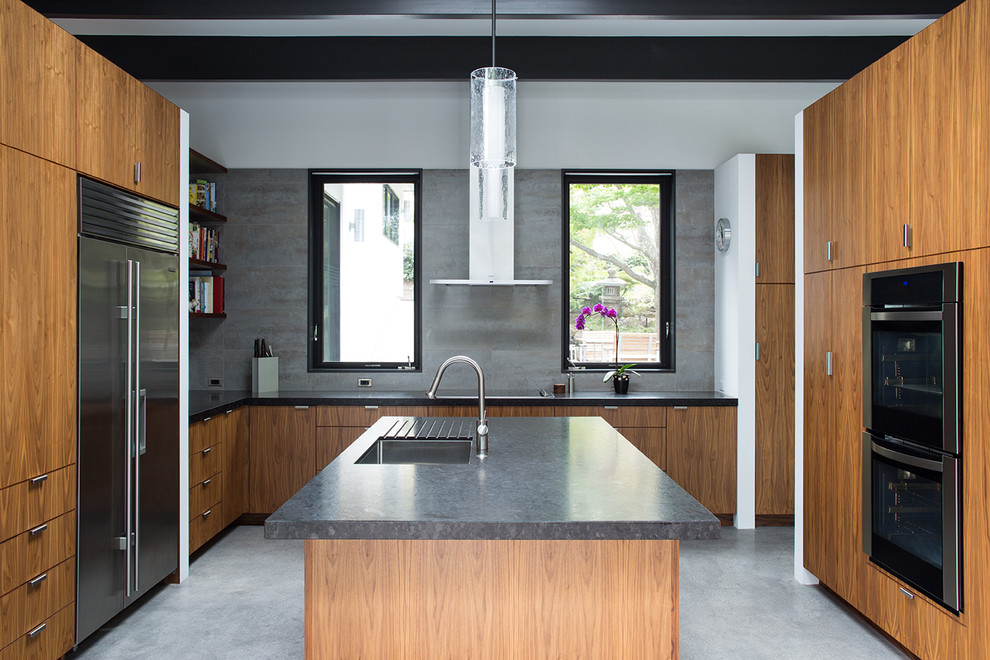 Bild på ett funkis kök, med en undermonterad diskho, släta luckor, skåp i mellenmörkt trä, grått stänkskydd, rostfria vitvaror, betonggolv och en köksö