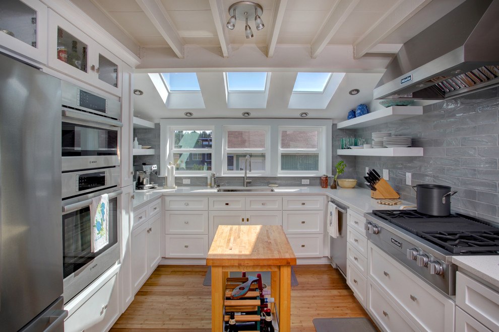 Foto på ett vintage u-kök, med en undermonterad diskho, skåp i shakerstil, vita skåp, grått stänkskydd, rostfria vitvaror, mellanmörkt trägolv, en köksö och brunt golv