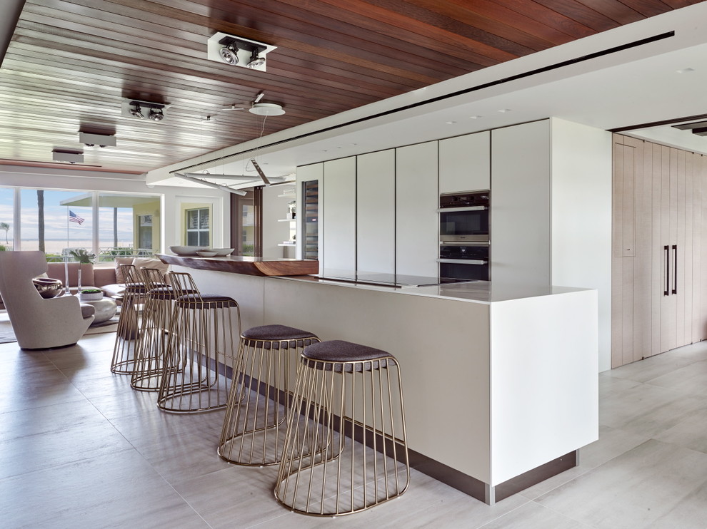Offene, Zweizeilige Moderne Küche mit flächenbündigen Schrankfronten, weißen Schränken, Küchengeräten aus Edelstahl, Kücheninsel, beigem Boden und weißer Arbeitsplatte in Miami