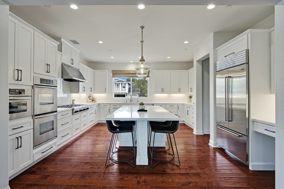 Inspiration for a large coastal u-shaped medium tone wood floor and brown floor open concept kitchen remodel in San Diego with an undermount sink, shaker cabinets, white cabinets, quartz countertops, white backsplash, ceramic backsplash, stainless steel appliances, an island and white countertops