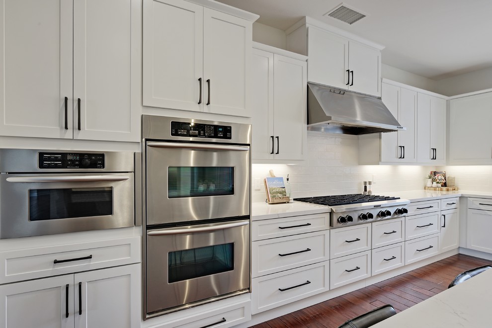 Foto på ett stort maritimt vit kök, med en undermonterad diskho, skåp i shakerstil, vita skåp, bänkskiva i kvarts, vitt stänkskydd, stänkskydd i keramik, rostfria vitvaror, mellanmörkt trägolv, en köksö och brunt golv