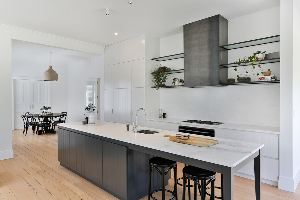 Inspiration for a mid-sized transitional galley light wood floor and brown floor open concept kitchen remodel in Auckland with white cabinets, marble countertops, white backsplash, marble backsplash, black appliances, an island, white countertops, an undermount sink and flat-panel cabinets