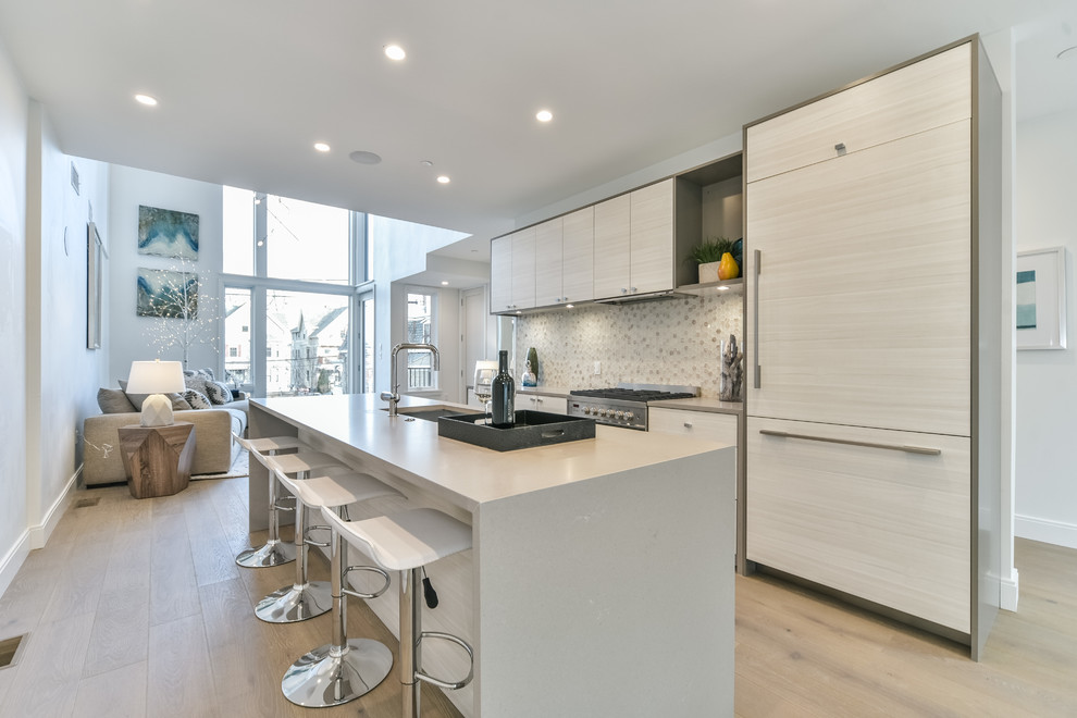 Example of a beach style light wood floor and beige floor open concept kitchen design in Boston with an undermount sink, flat-panel cabinets, beige cabinets, multicolored backsplash, paneled appliances, an island and gray countertops