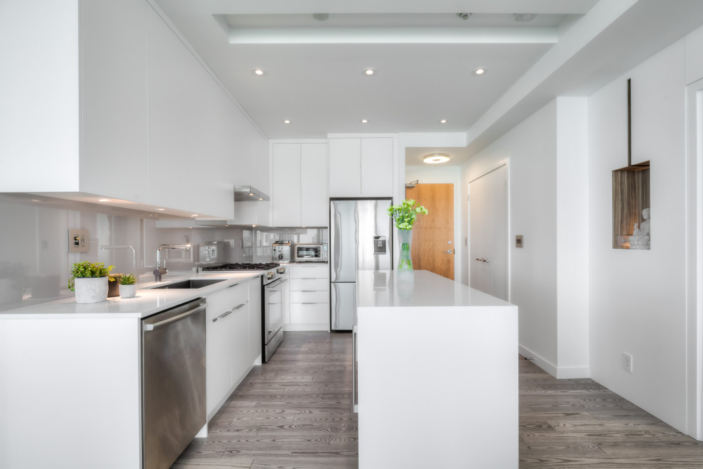 Small contemporary galley kitchen/diner in Toronto with a submerged sink, flat-panel cabinets, white cabinets, engineered stone countertops, grey splashback, glass sheet splashback, stainless steel appliances, medium hardwood flooring, an island, grey floors, white worktops and a coffered ceiling.