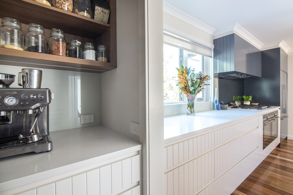 Foto på ett stort funkis vit kök, med en dubbel diskho, vita skåp, bänkskiva i kvarts, rostfria vitvaror, mellanmörkt trägolv, en köksö, brunt golv, vitt stänkskydd och glaspanel som stänkskydd