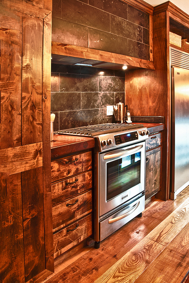 Cette image montre une cuisine américaine parallèle rustique en bois brun avec un évier encastré, un placard à porte plane, un plan de travail en bois, une crédence noire, une crédence en carrelage de pierre et un électroménager en acier inoxydable.