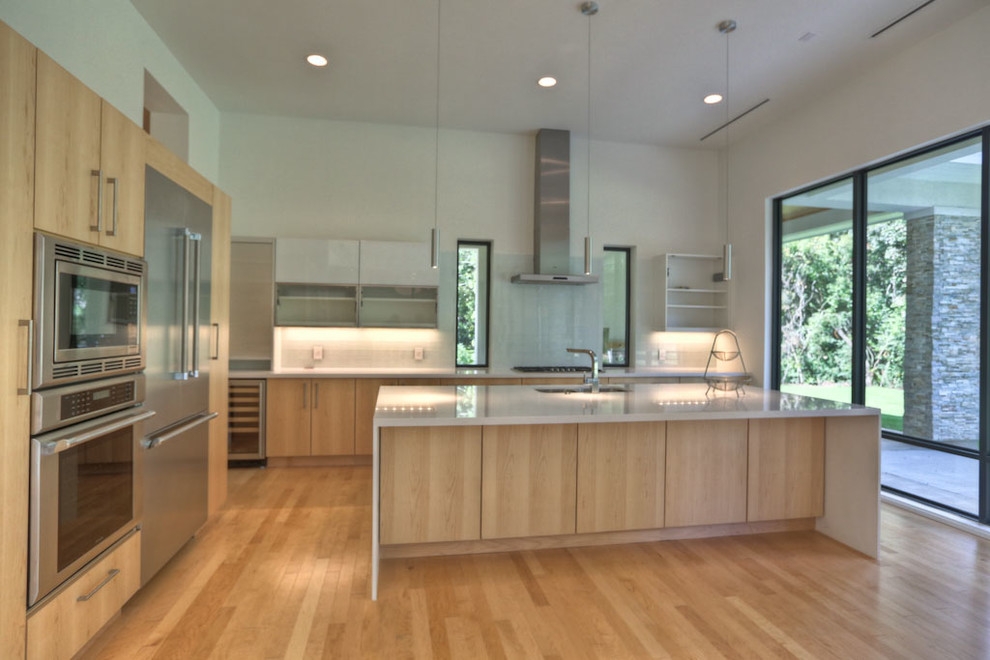 Photo of a large contemporary l-shaped kitchen/diner in Houston with a double-bowl sink, flat-panel cabinets, light wood cabinets, engineered stone countertops, white splashback, ceramic splashback, stainless steel appliances, light hardwood flooring and an island.