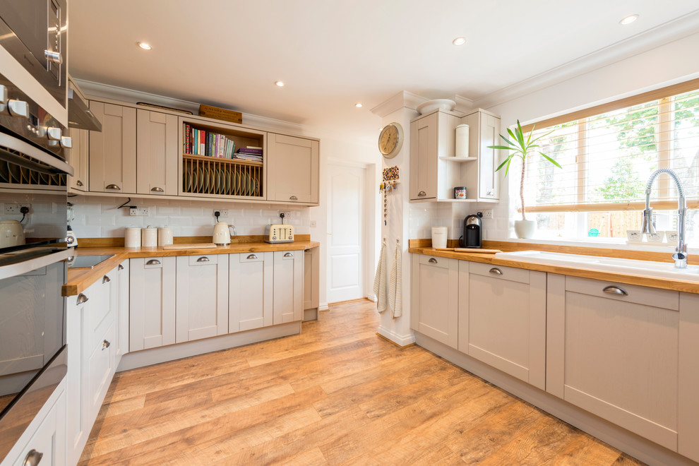 Inspiration for a mid-sized country laminate floor and brown floor enclosed kitchen remodel in Dorset with a double-bowl sink, shaker cabinets, beige cabinets, wood countertops, stainless steel appliances, white backsplash and subway tile backsplash